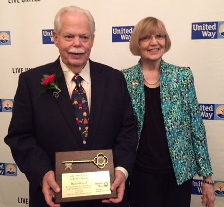 Man holds award with key; woman stands beside him. Both smile at an event with a United Way backdrop.
