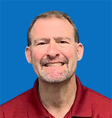Man with short brown hair and beard smiles, wearing a red polo, against a teal background.