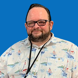 Man with glasses and beard wearing a boat-print shirt smiles in front of a blue wall.