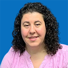 Woman with curly dark hair smiles, wearing a pink shirt, in front of a blue background.