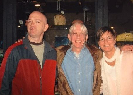 Three people smiling, posing for photo. One bald man in red jacket, man in middle, and woman in pink. Indoor setting.