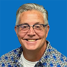 Man with gray hair and glasses smiles, wearing a colorful shirt. Blue background.