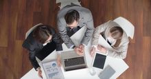 A group of business people are sitting around a table looking at a laptop.