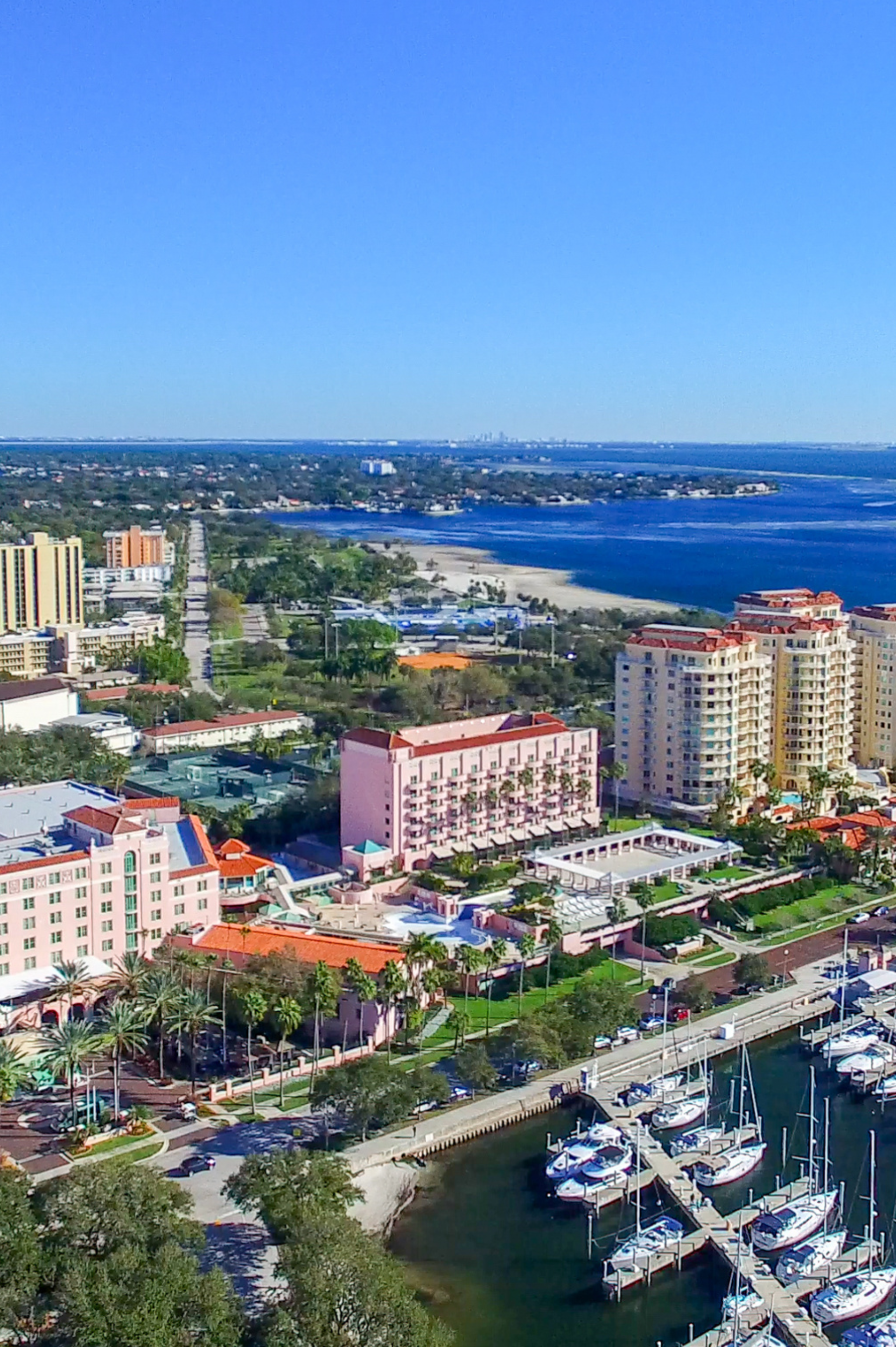 house painters dunedin - Aerial view of a coastal city featuring colorful buildings, palm trees, a marina with boats, and a beach under a clear blue sky.