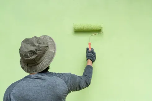 house painters dunedin - A person wearing a hat and gloves is painting a light green wall with a roller, focused on their task.