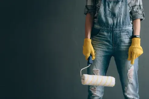 house painters dunedin - A person in denim overalls and yellow gloves holds a paint roller against a textured gray wall, ready for a painting project.