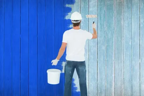 house painters dunedin - A painter in a white helmet applies blue paint to a wooden wall, holding a paint roller and bucket.