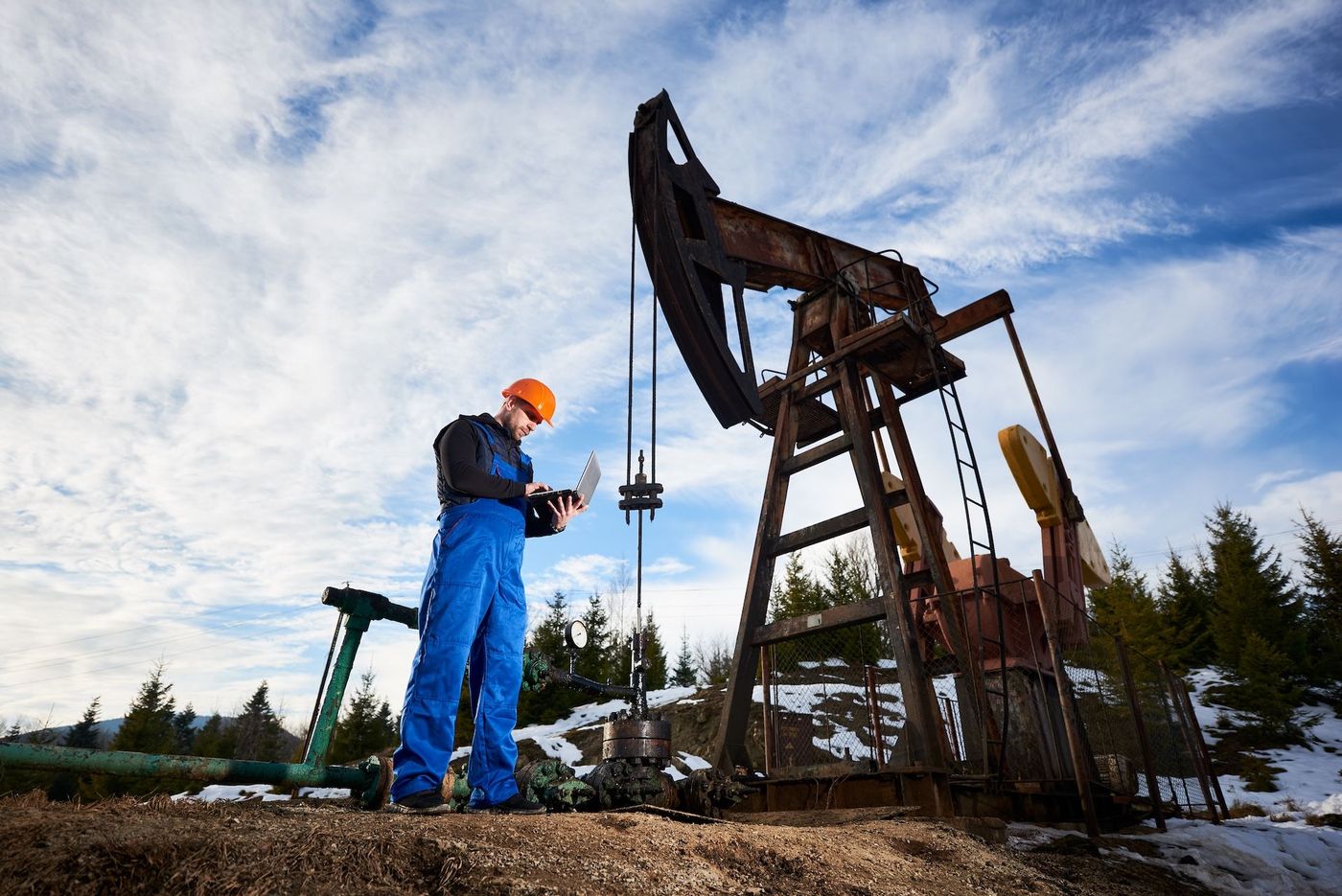 A man in with a computer and hard hat is entering data about the oil pump behind him.