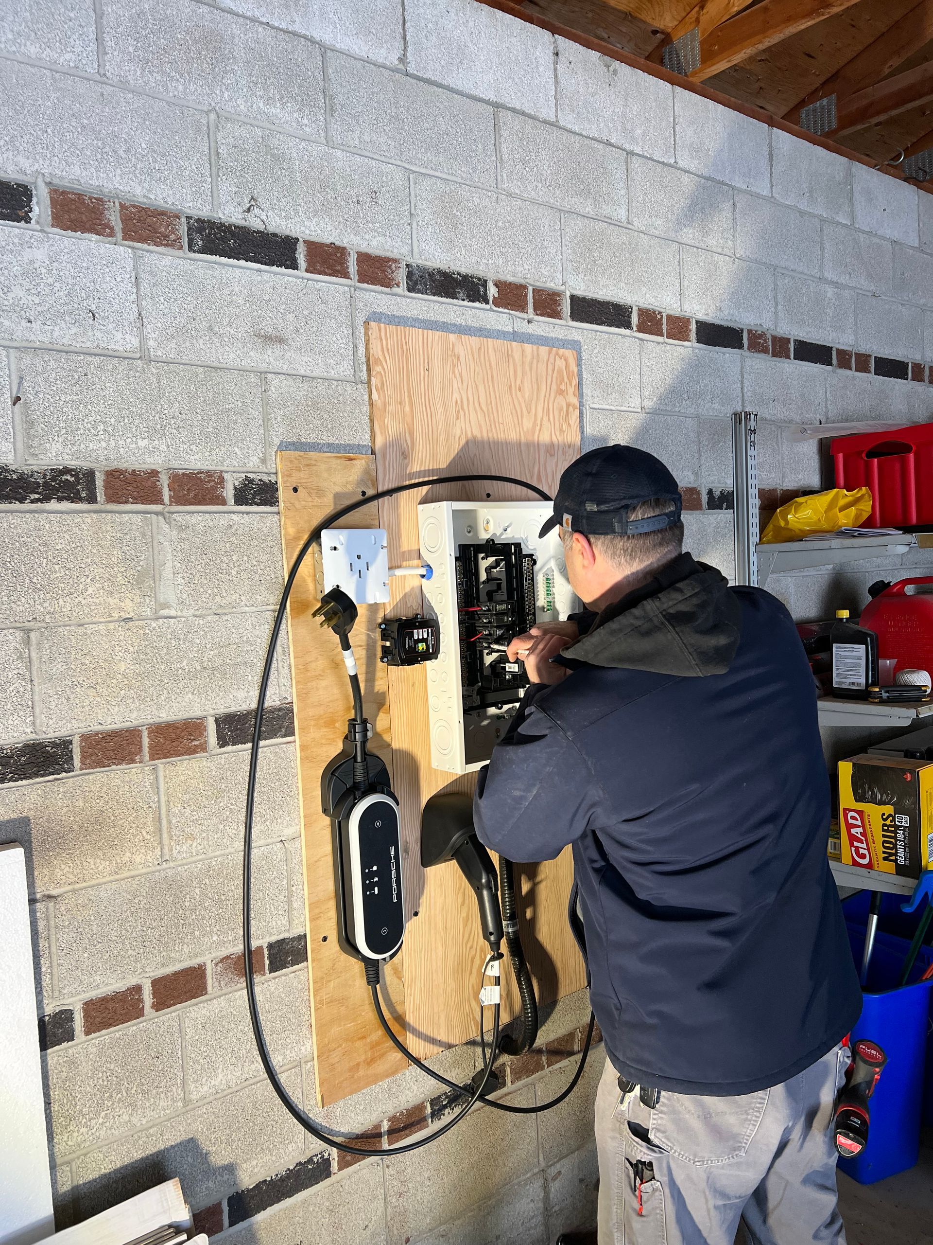 Man installing electrical equipment in garage; black and grey wiring on a wooden board, concrete block wall.