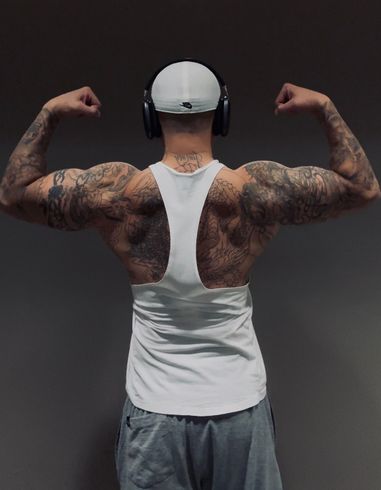 Man flexing biceps, wearing a white tank top and gray shorts. Tattooed arms and back. Gray background.