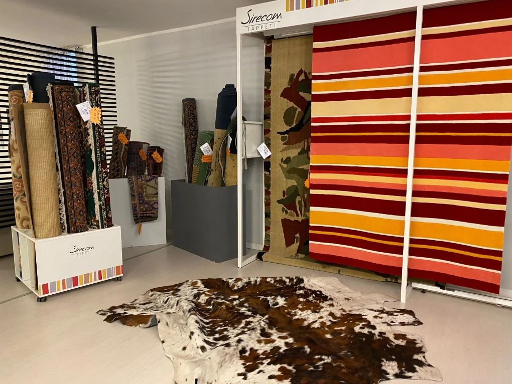 Esposizione di tappeti in un negozio, con vari tappeti arrotolati e appesi. Un tappeto in pelle di mucca è appoggiato sul pavimento.