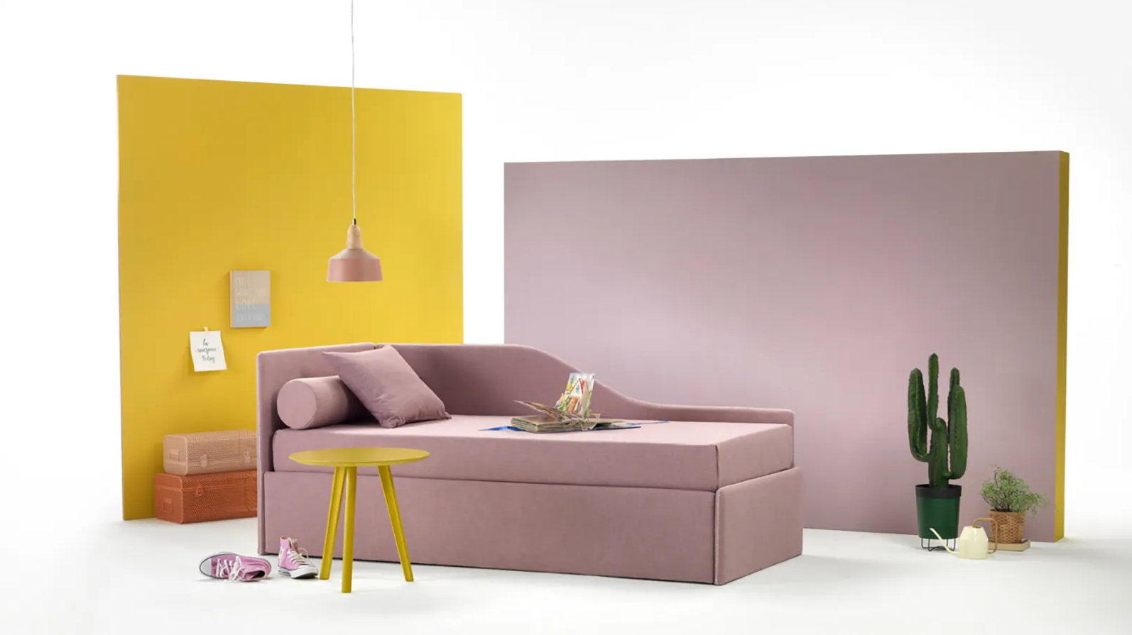 Divano letto rosa con cuscini decorativi, tavolino giallo, su sfondo giallo e rosa, cactus, lampada a sospensione.