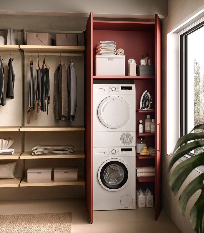 Lavanderia con lavatrice/asciugatrice impilate, scaffalature e vestiti appesi; mobili color bordeaux, pareti neutre.