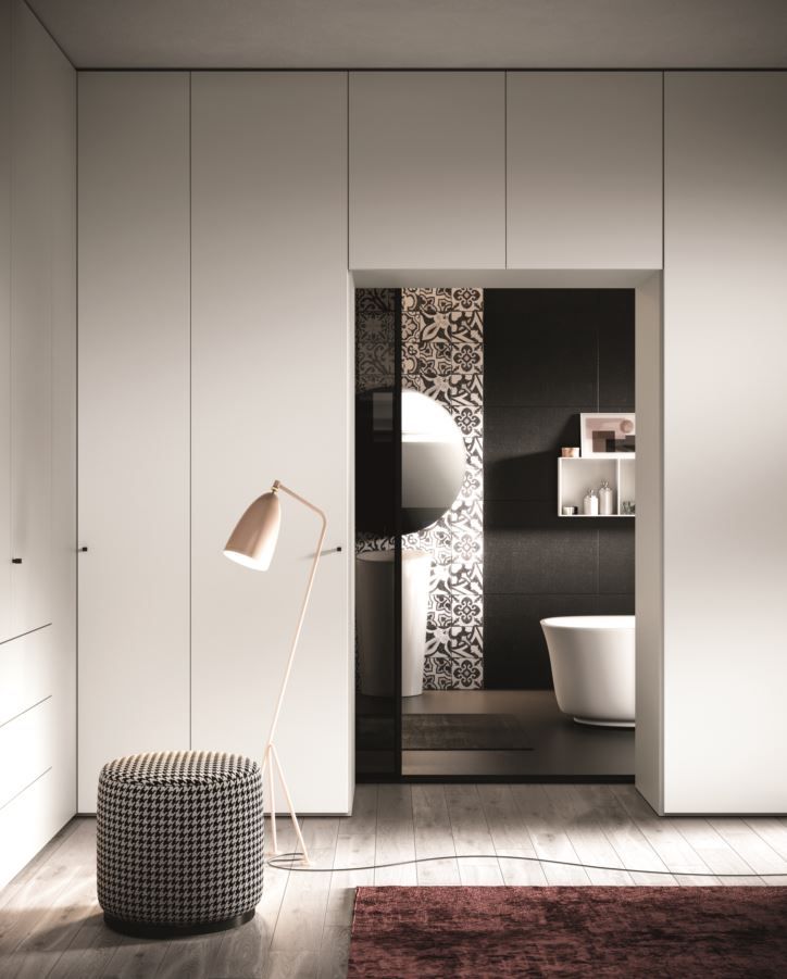 Camera da letto moderna con armadio bianco, che conduce a un bagno con specchio circolare e vasca.