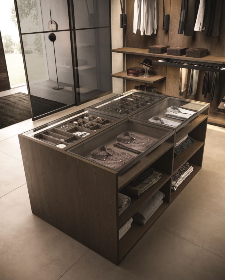 Isola armadio in legno scuro con piano espositivo in vetro, per riporre vestiti e accessori. I ripiani contengono oggetti piegati.