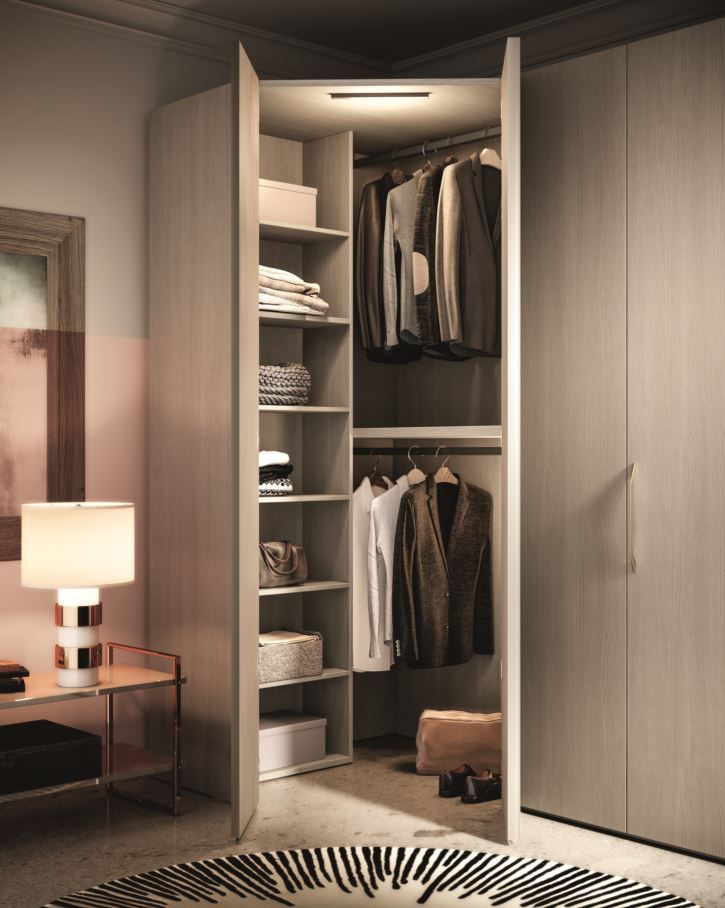 Cabina armadio con ante aperte, che mostra vestiti, mensole e un tappeto. Dominano i toni chiari del legno.