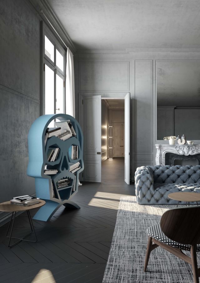 Libreria azzurra a forma di teschio piena di libri, in un soggiorno nei toni del grigio con un divano blu trapuntato.