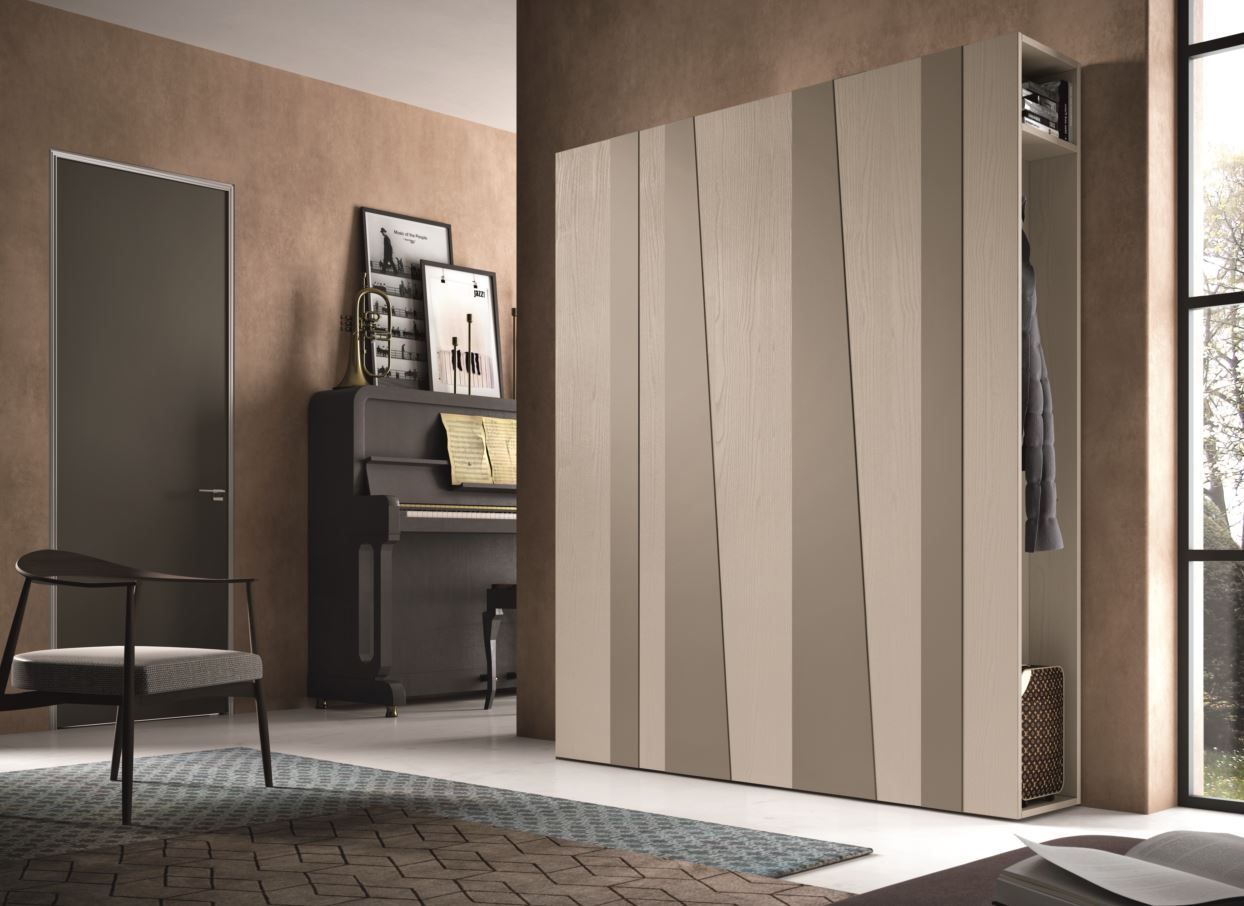 Armadio moderno con pannelli in legno chiaro angolati, che si aprono per rivelare i vestiti appesi, vicino al pianoforte e alla sedia.