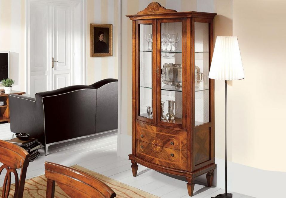 Vetrina in legno con ante in vetro, contenente posate. Lampada alta a destra, stanza con divano e opere d'arte visibili.