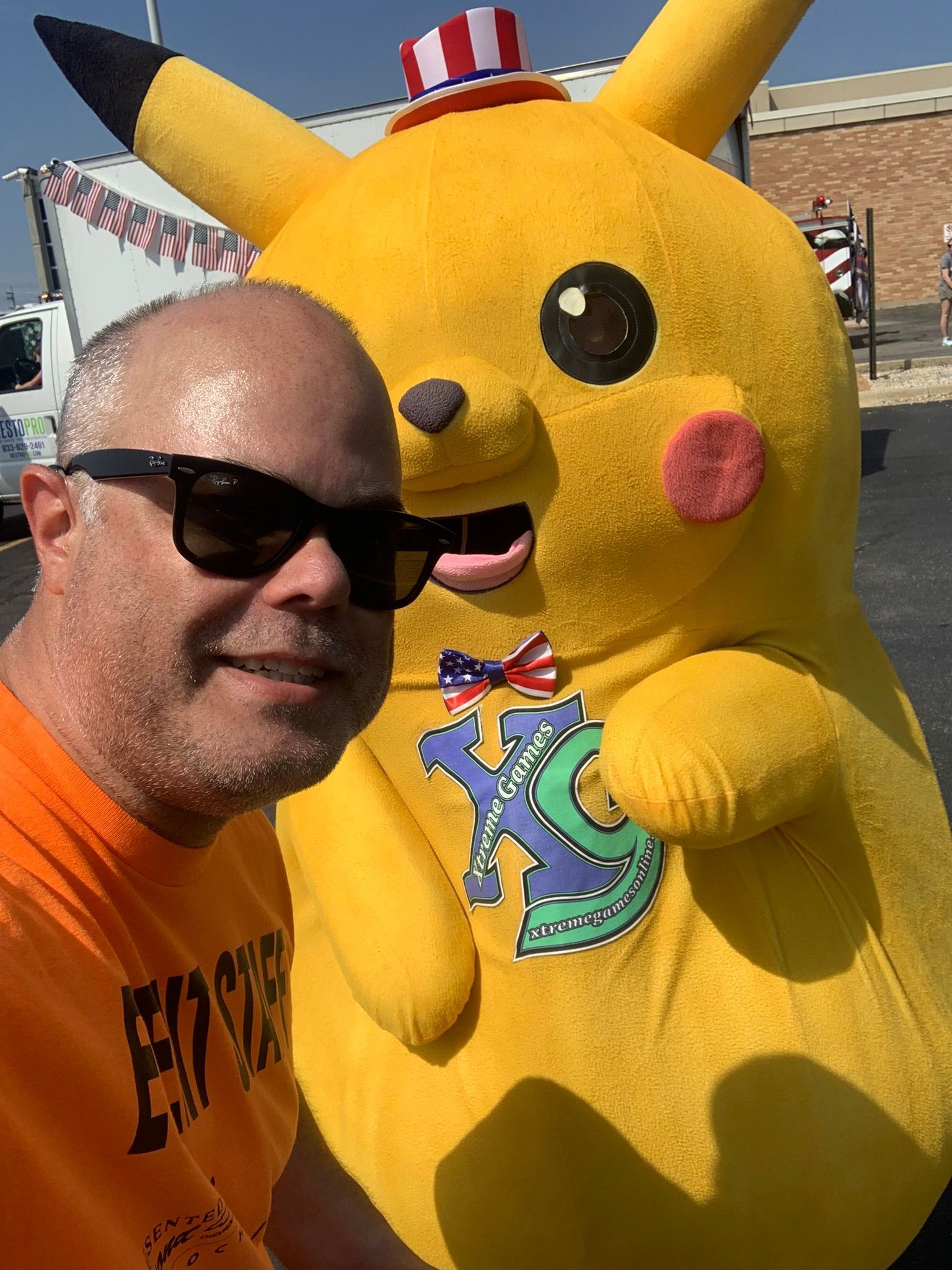 Man in sunglasses takes a selfie with a Pikachu in a patriotic hat and bow tie, outdoors.