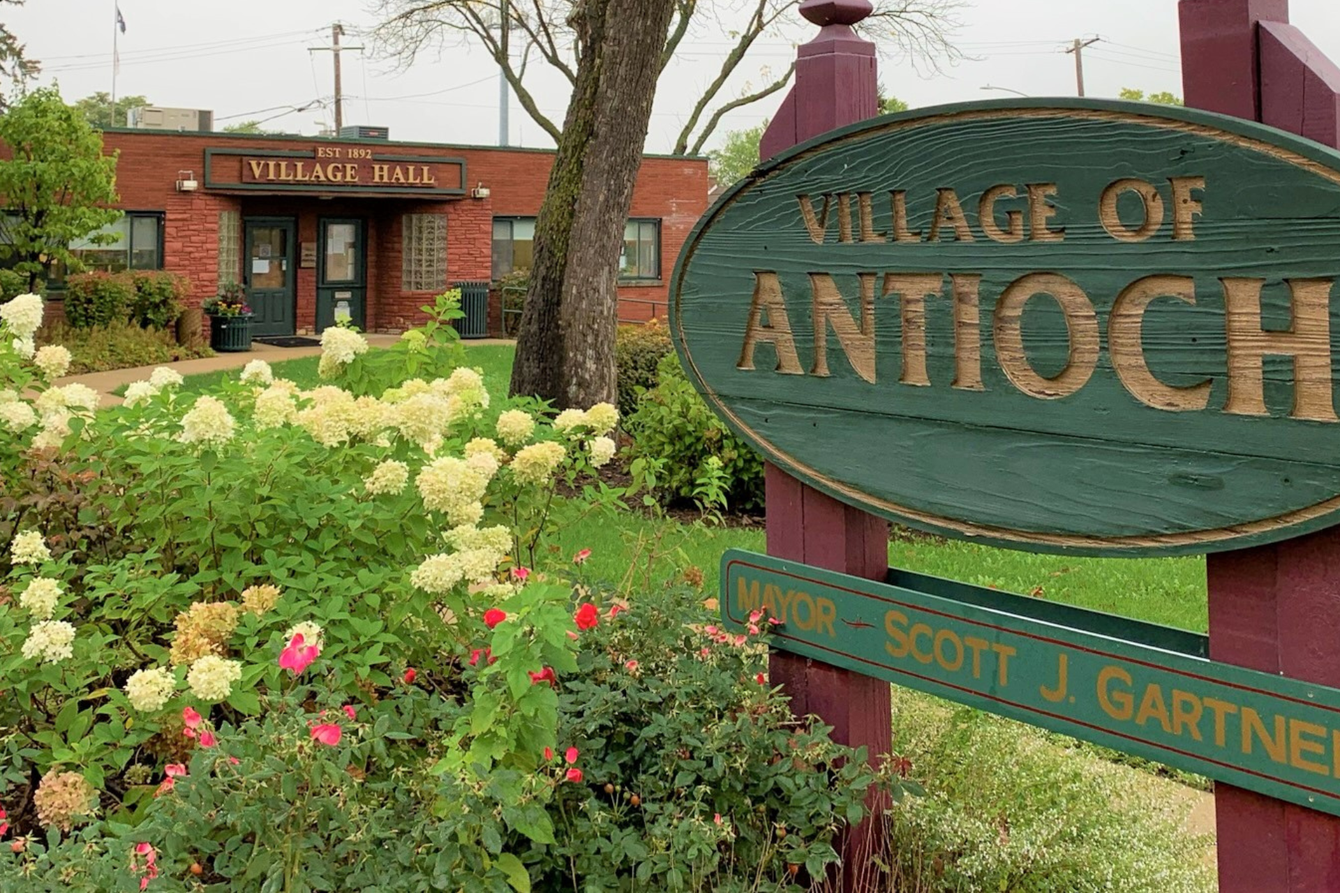 Village of Antioch sign with Village Hall in the background, surrounded by flowers and greenery.