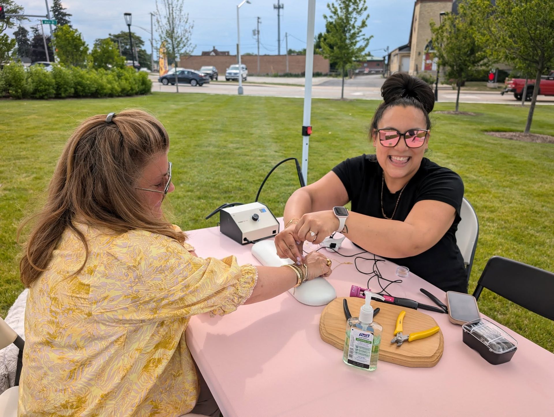 Two women at a pink table making jewelry outdoors. One is smiling and working on the other's wrist.