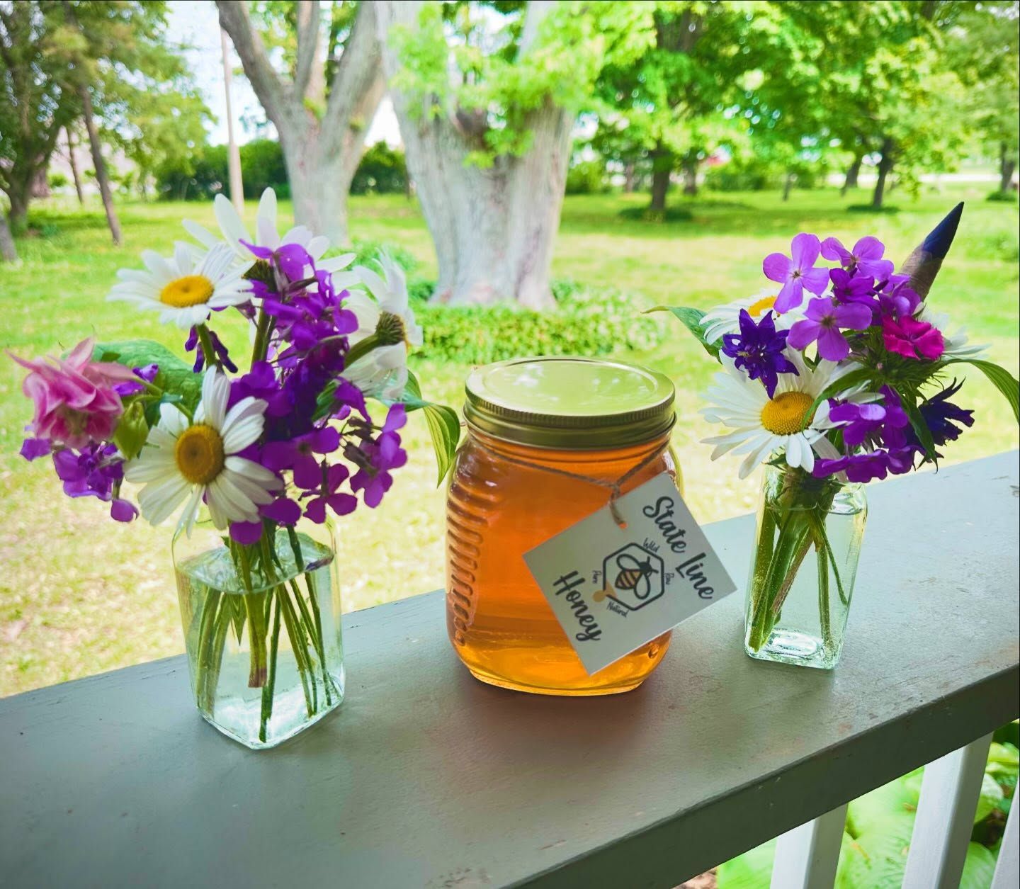 Jar of honey with a label, flanked by small flower bouquets on a porch railing with a grassy backdrop.