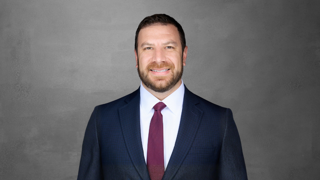 Man in navy suit, white shirt, and maroon tie smiles at the camera against a gray backdrop.