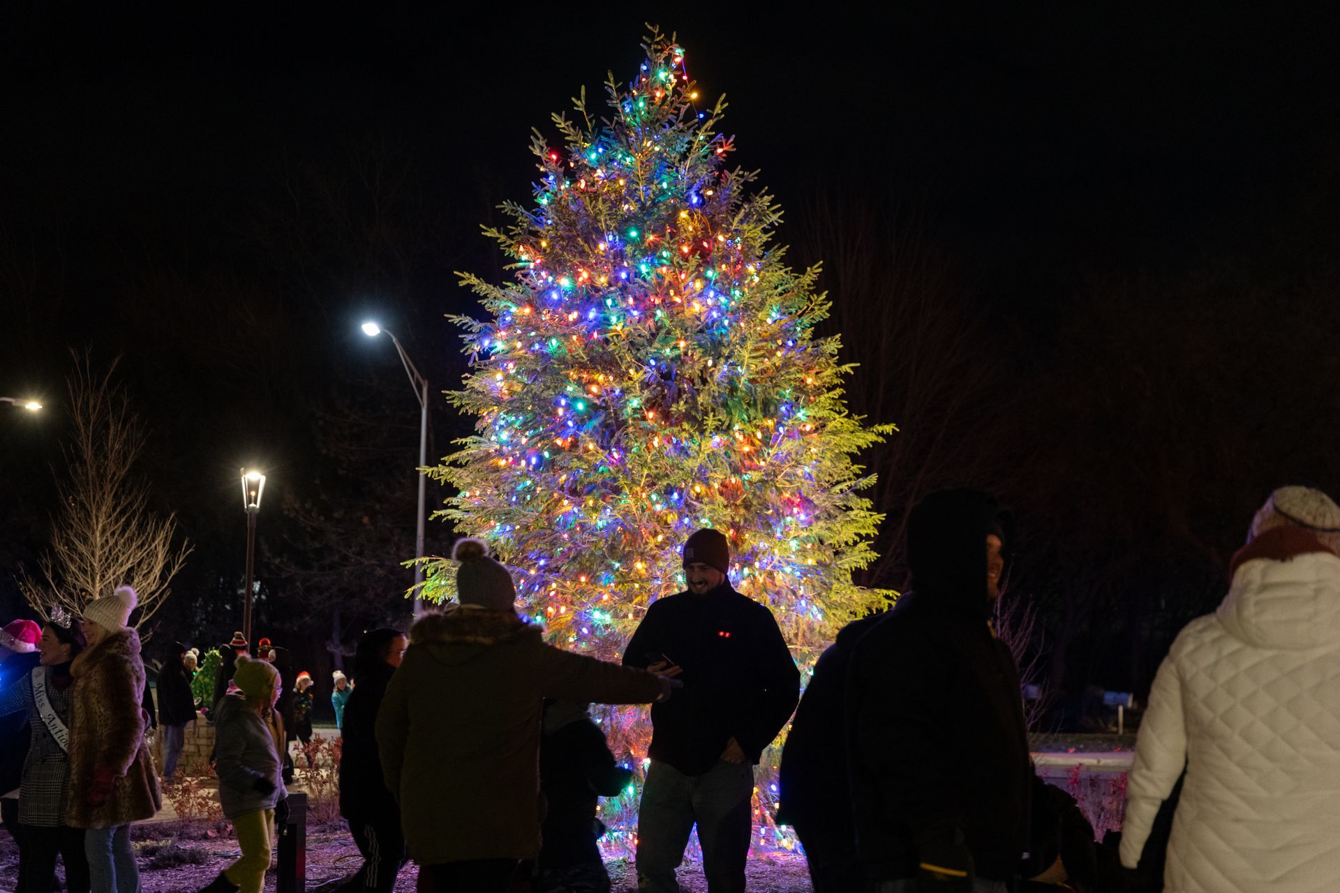 People gathered around a large Christmas tree lit with colorful lights at night.