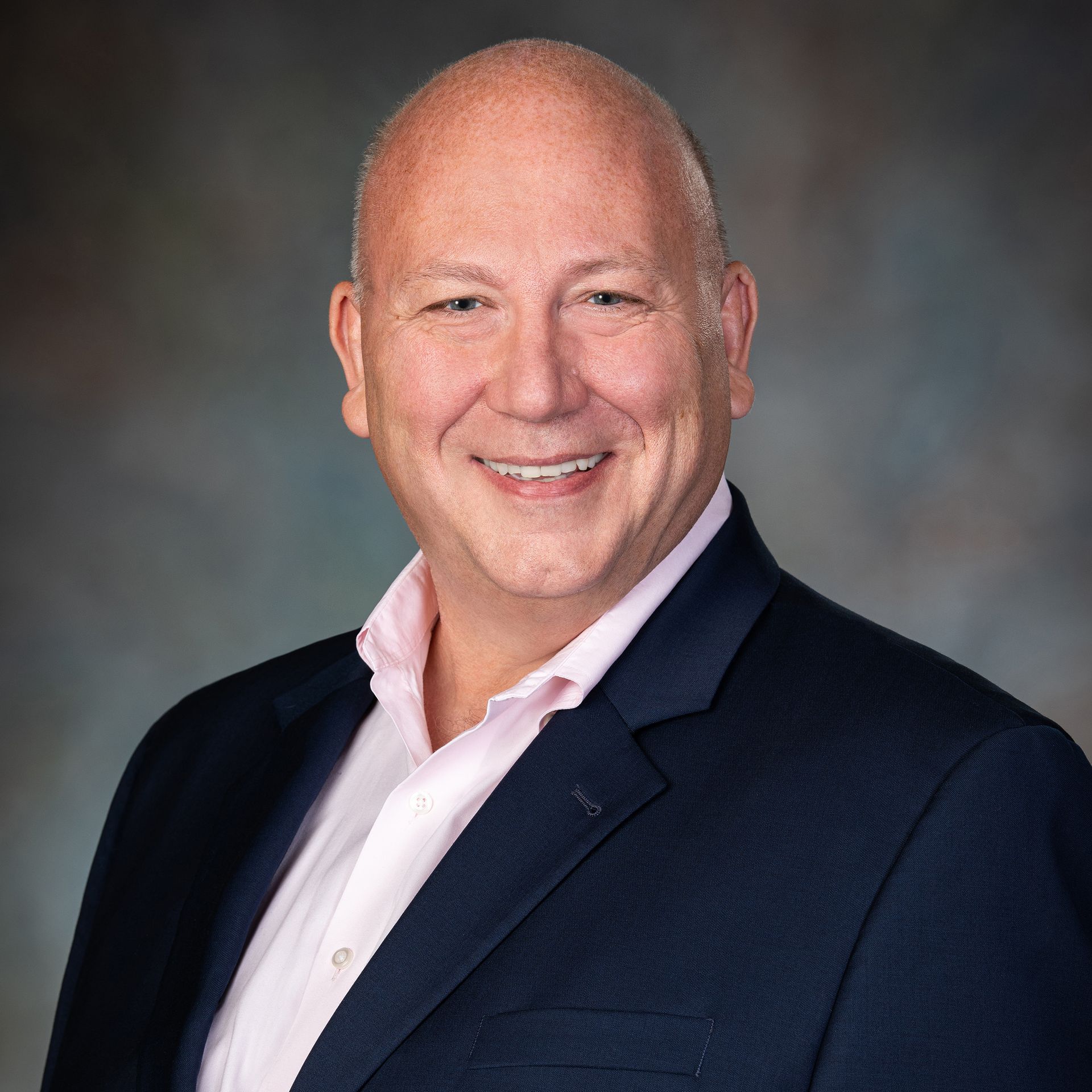 Smiling bald man in a blue blazer and pink shirt. Background is a neutral gray-blue gradient.
