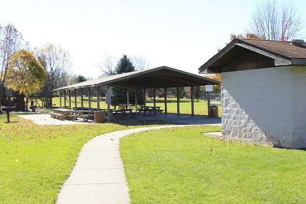 A paved path leads to a park shelter with picnic tables. Green grass, trees, and a bright sky are visible.