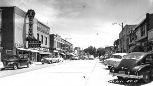 Black and white photo of a street lined with old buildings, cars parked on both sides; a theater marquee is visible.