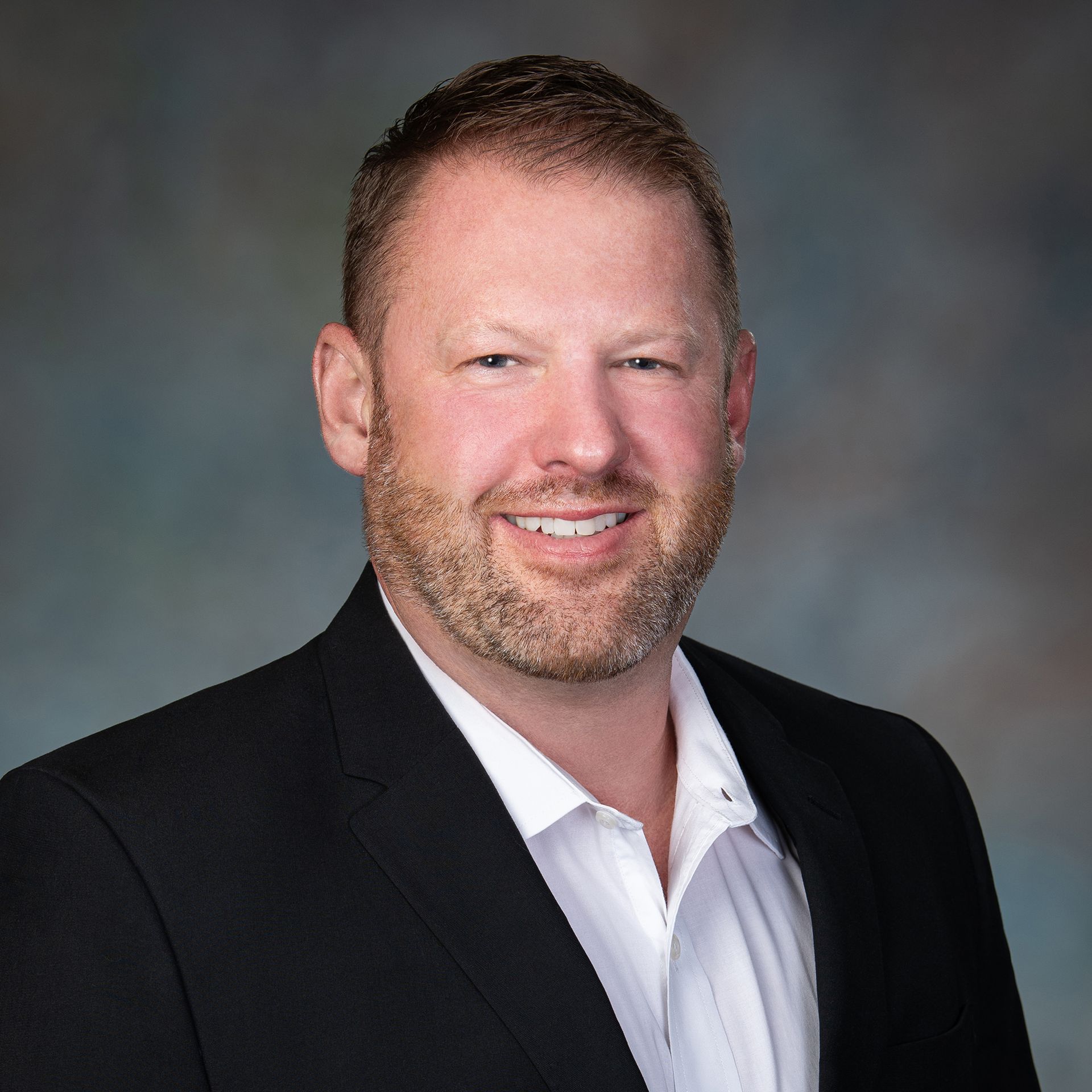 Man in a black suit jacket and white shirt smiling. Against a blurred background.