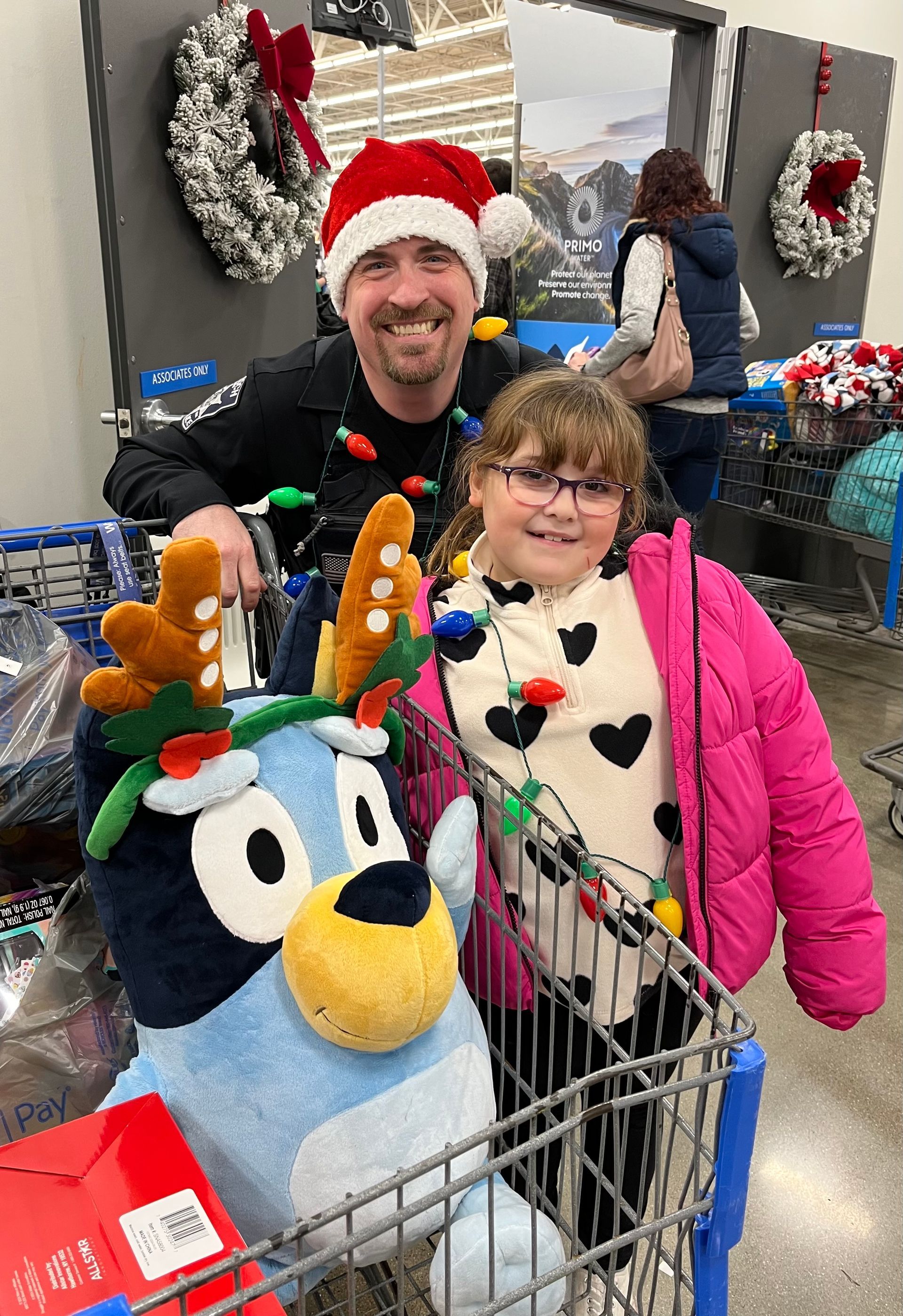 A father and daughter with a Bluey plush in a shopping cart at a store during the holidays, wearing Christmas lights and hats.