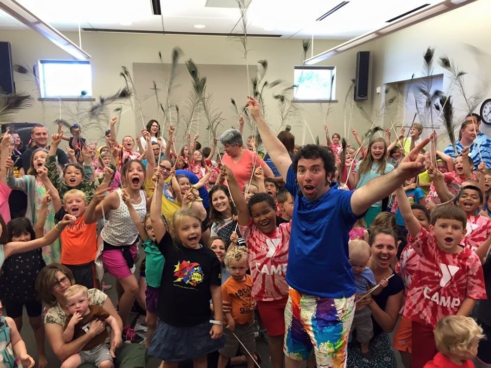 Group of children and adults in a room, arms raised in excitement. Tie-dye shirts, peacock feathers, bright expressions.