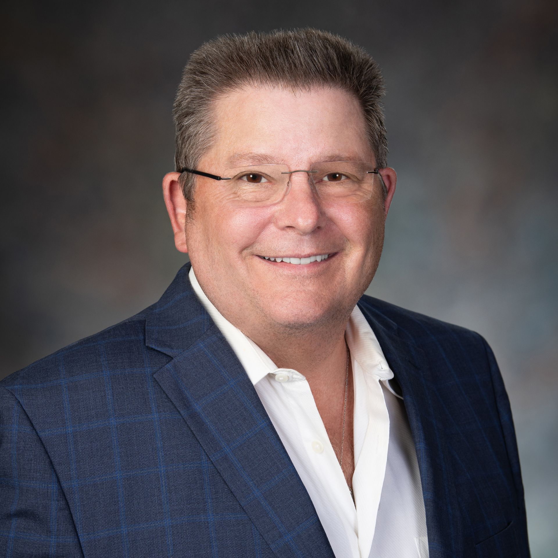 Smiling man in glasses, blazer, and open-collared shirt. Studio portrait with neutral background.