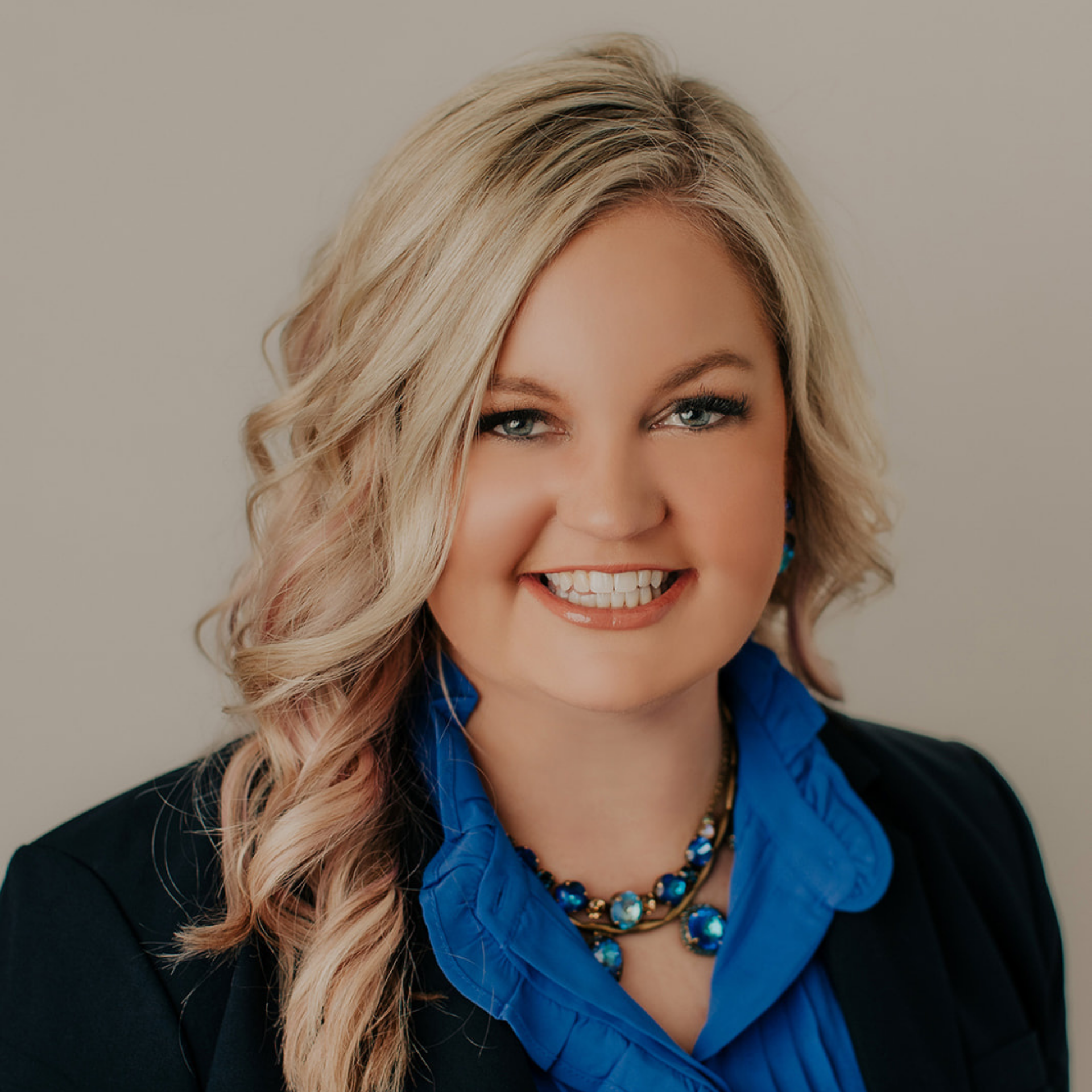 Blonde woman smiling, wearing a blue blouse and necklace, blazer.