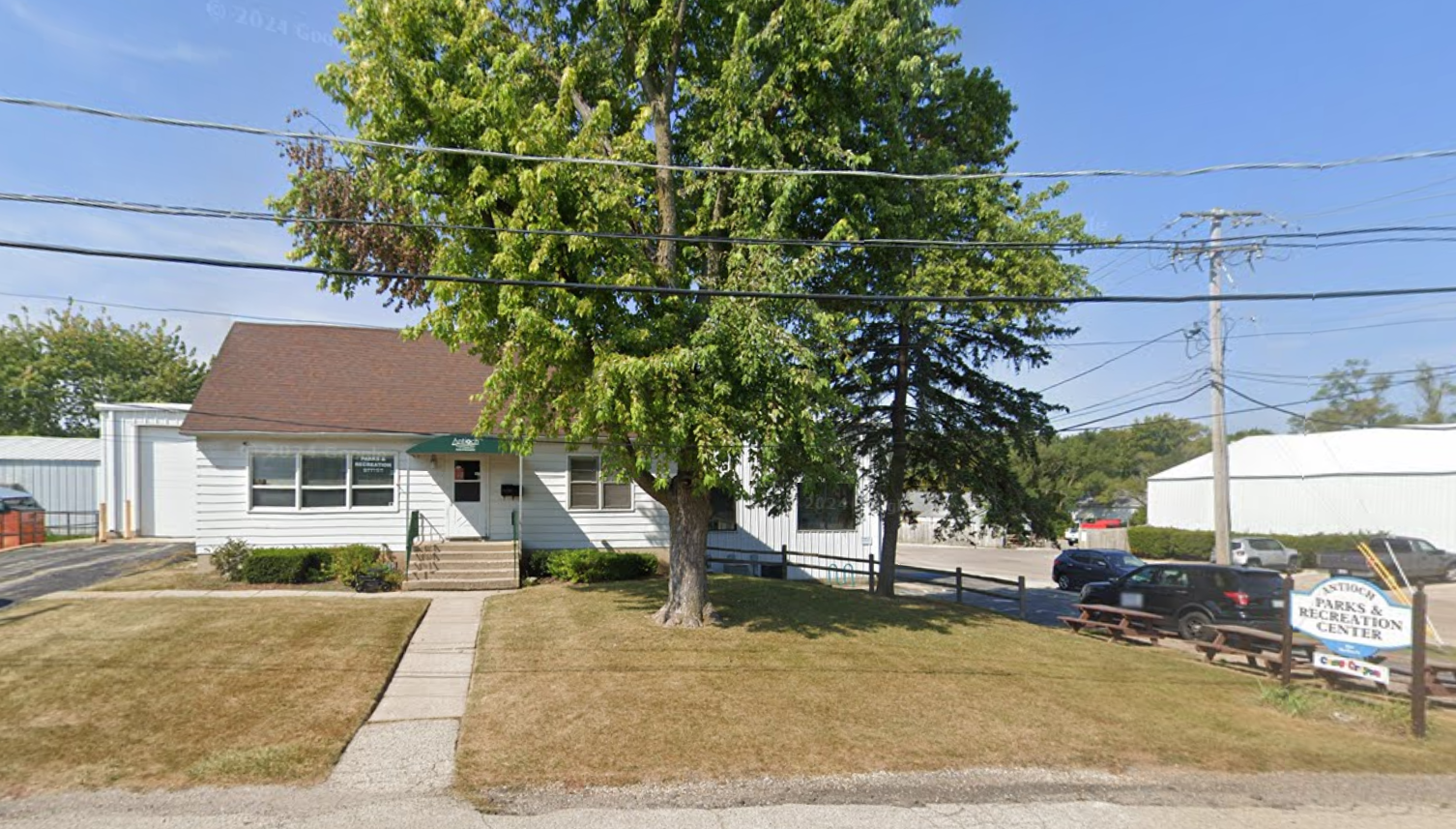 White house with brown roof, tree in front, sign on the right side, and short cut brown grass.