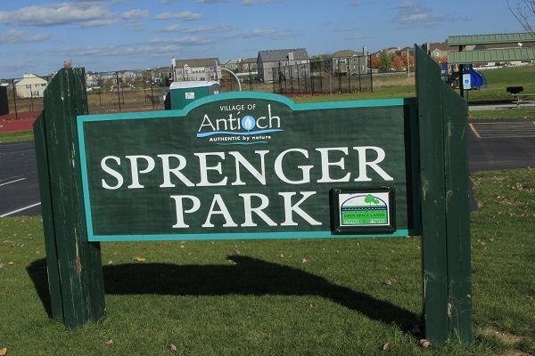 Sign for Sprenger Park in Antioch, Illinois, with a green and teal color scheme, set against a grassy field under a sunny sky.
