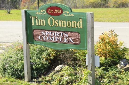 Sign for Tim Osmond Sports Complex, green and brown, established 2018, near a road, with foliage.