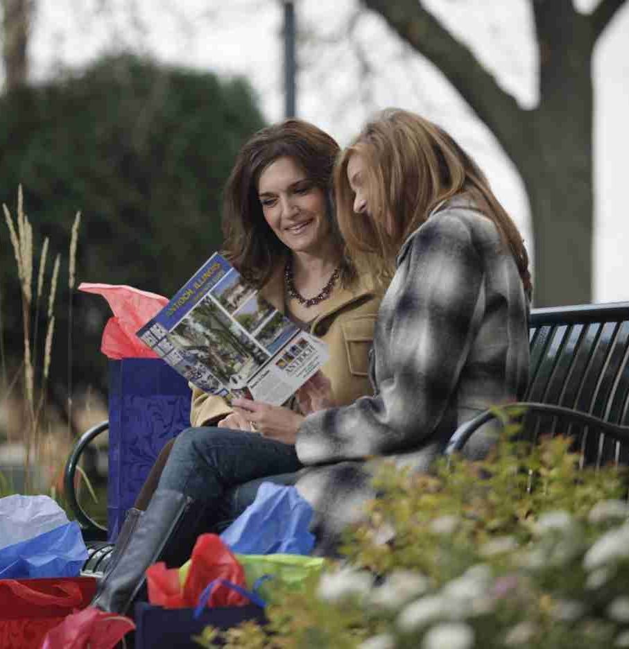 Two women seated on a park bench, looking at a brochure. They are surrounded by shopping bags.