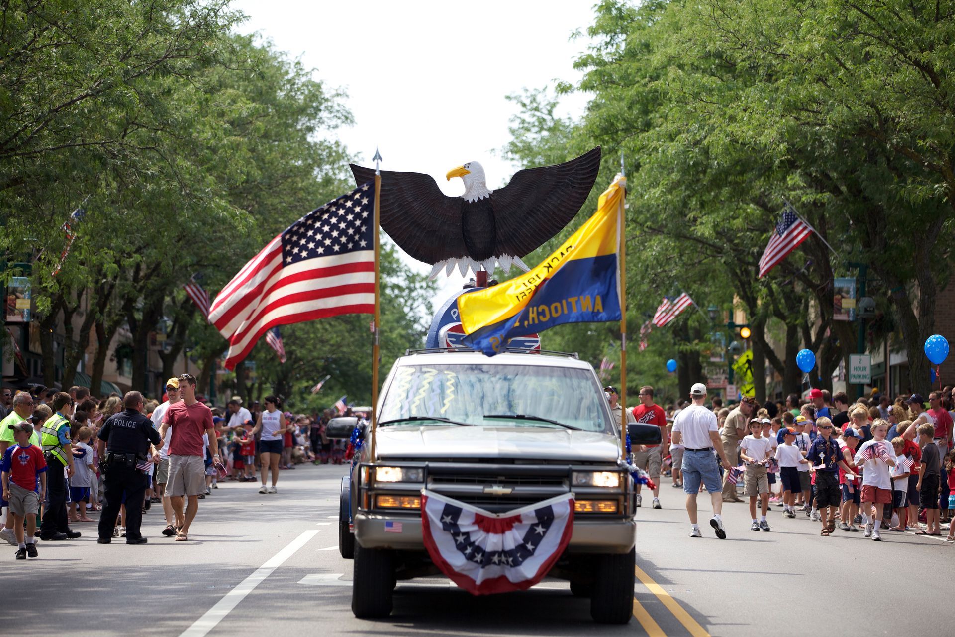 A parade with an American flag, large eagle, and Chevrolet van driving down a street lined with spectators.