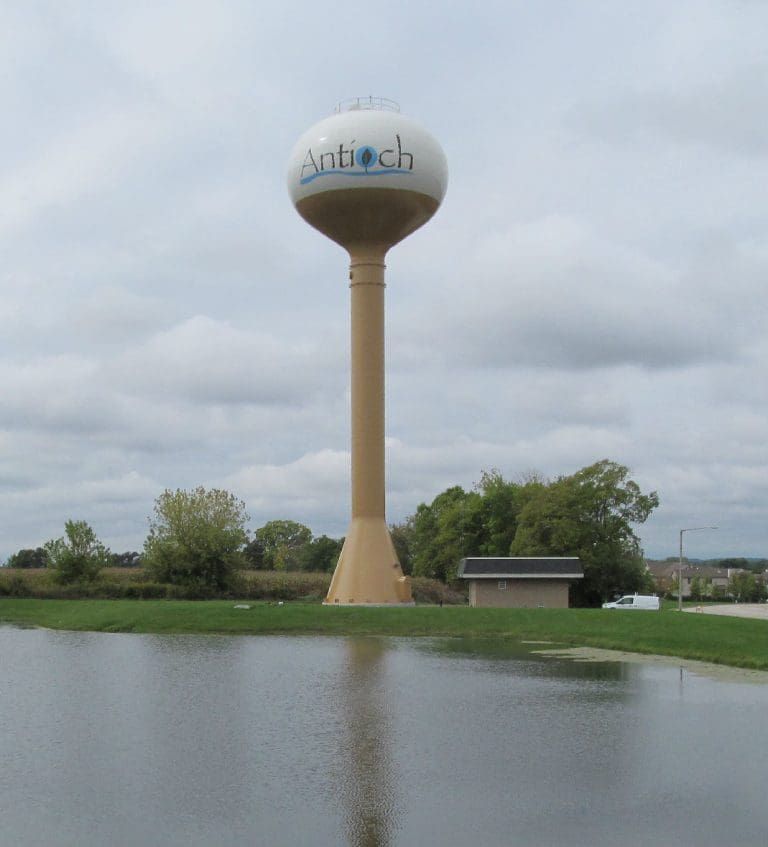 Water tower in Antioch, IL. Beige with blue logo, tall, over a pond, cloudy sky.