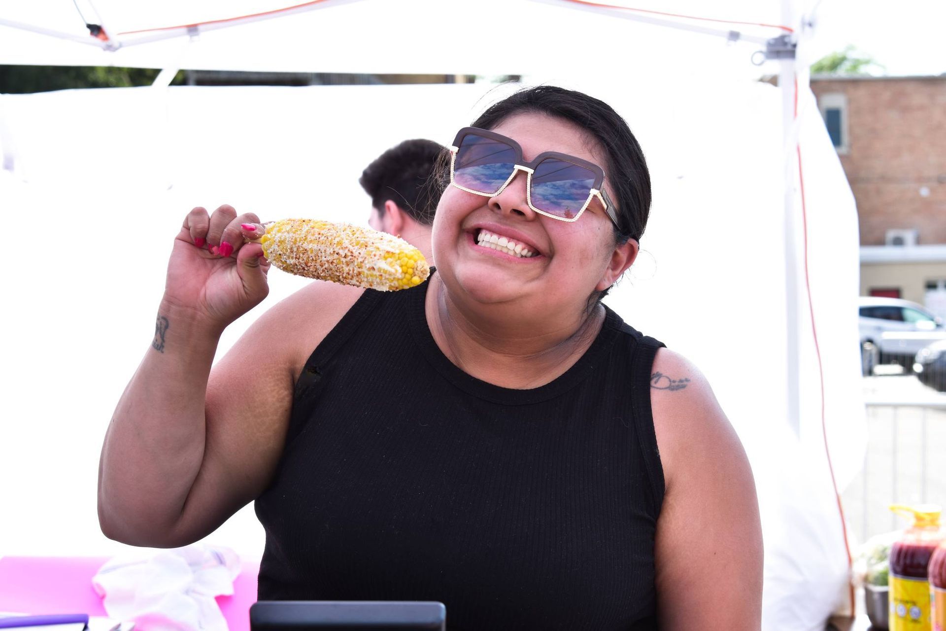 Woman smiling, holding corn on the cob, wearing sunglasses and black tank top. Outdoors at a market.