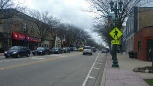 Street scene: cars on road, pedestrian crossing sign, shops, bare trees, sidewalk.