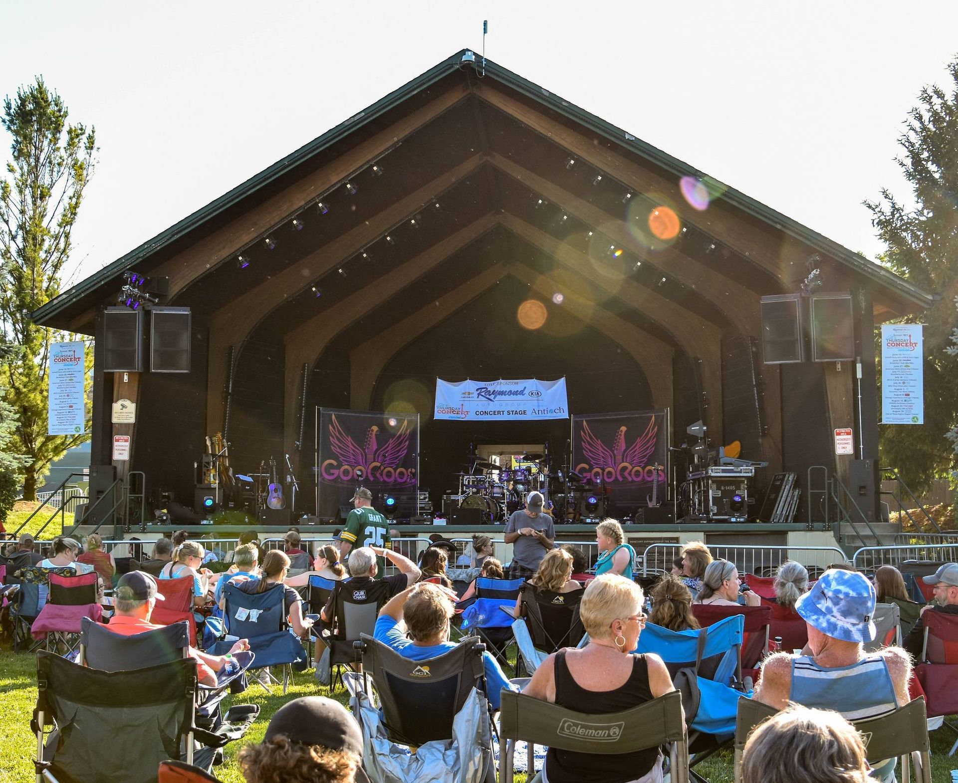 Outdoor concert: band performing on stage, audience seated in chairs, sunny day.