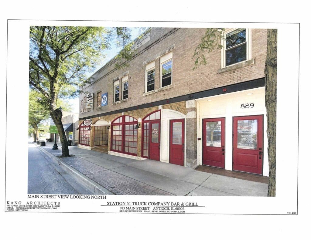 Brick building with red doors on a sidewalk. Trees line the street.