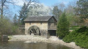 Wooden mill with a water wheel next to a pond, trees in the background.