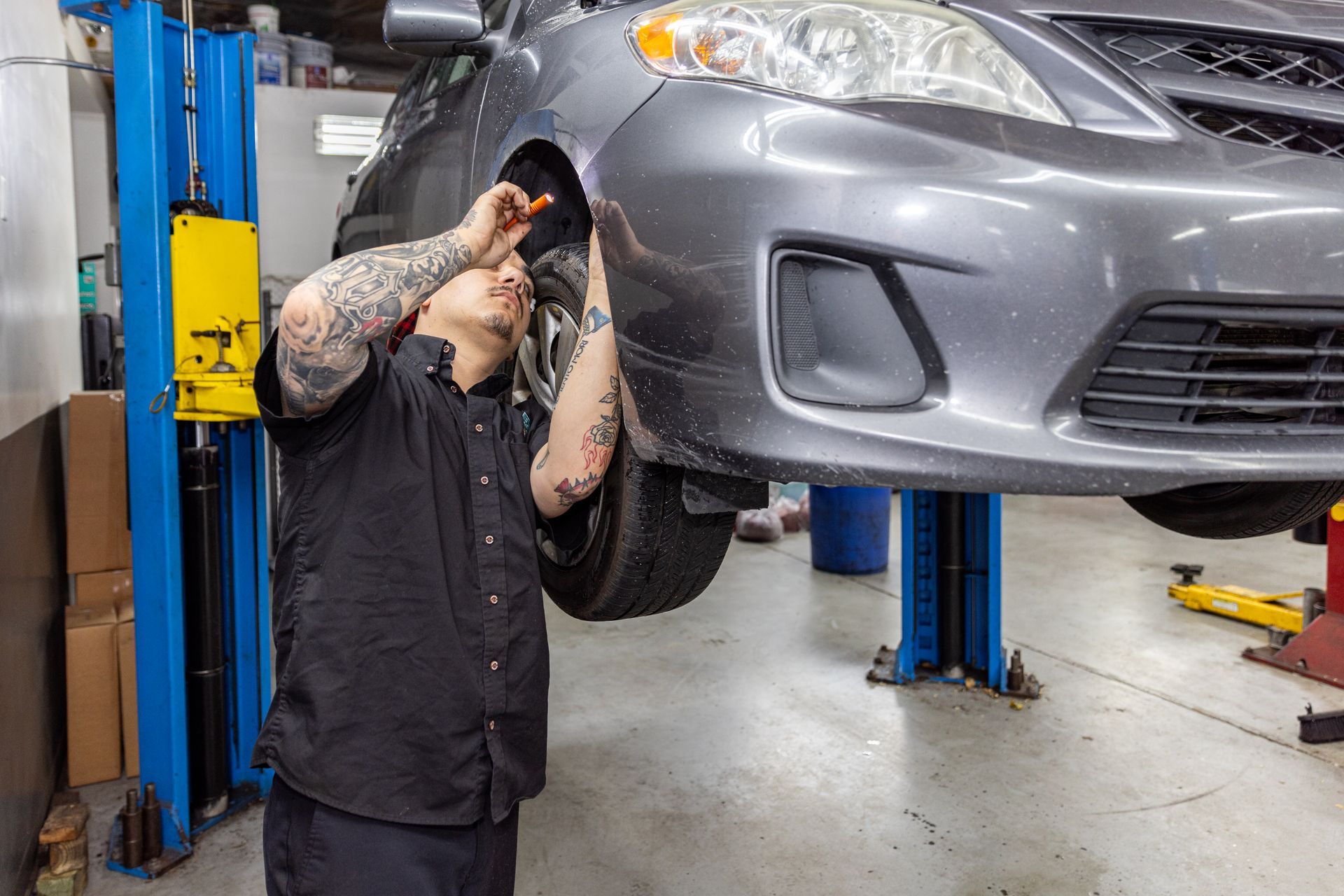 Mechanic working on a gray car raised on a lift in a garage. He is wearing a black shirt and has tattoos.