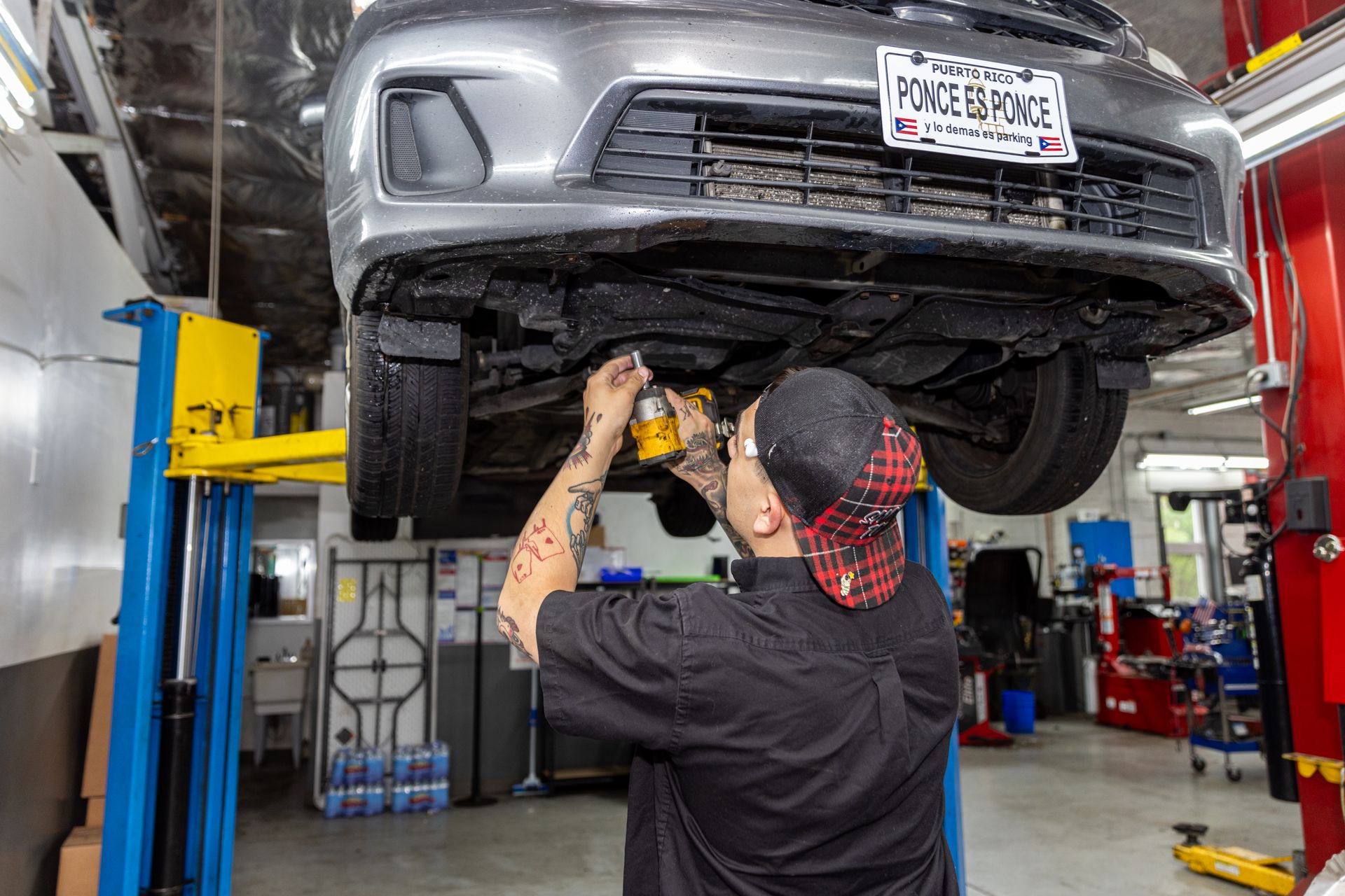 Mechanic working under a gray car lifted on a hydraulic lift in an auto repair shop.
